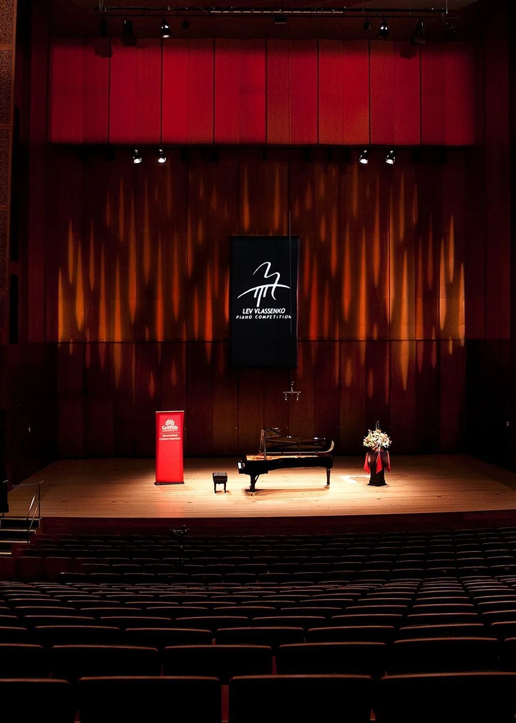 Lev Vlassenko Piano Competition - Empty stage with red drapes, piano, podium, and flower arrangement under warm stage lights