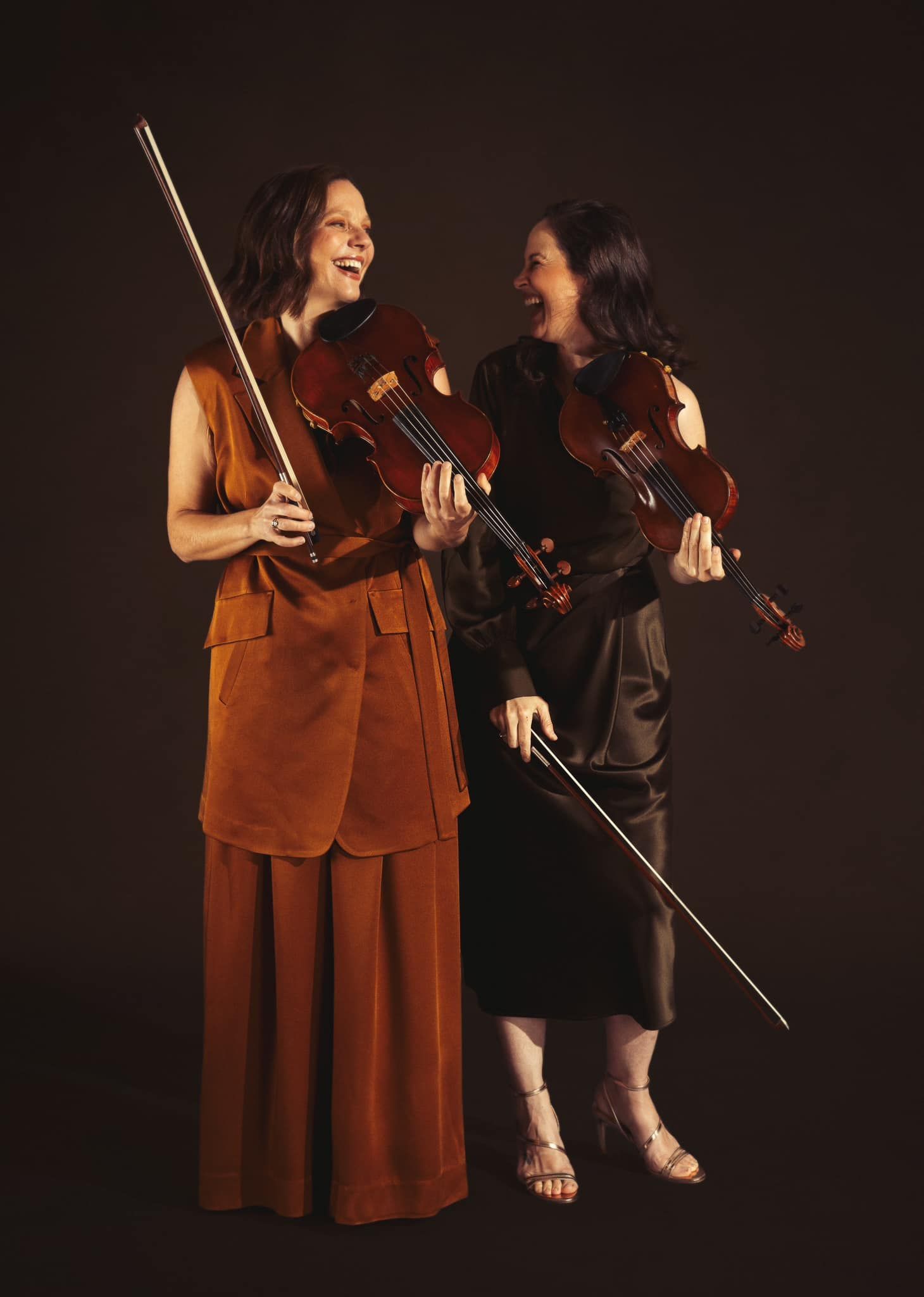 Queensland Symphony Orchestra - Two musicians in warm brown outfits holding string instruments on a dark stage, smiling at each other