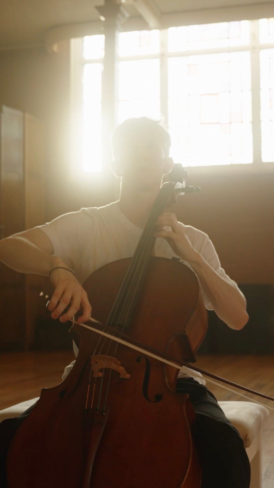 Queensland Youth Orchestras - Person playing a cello in a sunlit room at the Old Museum Building by a bright window.