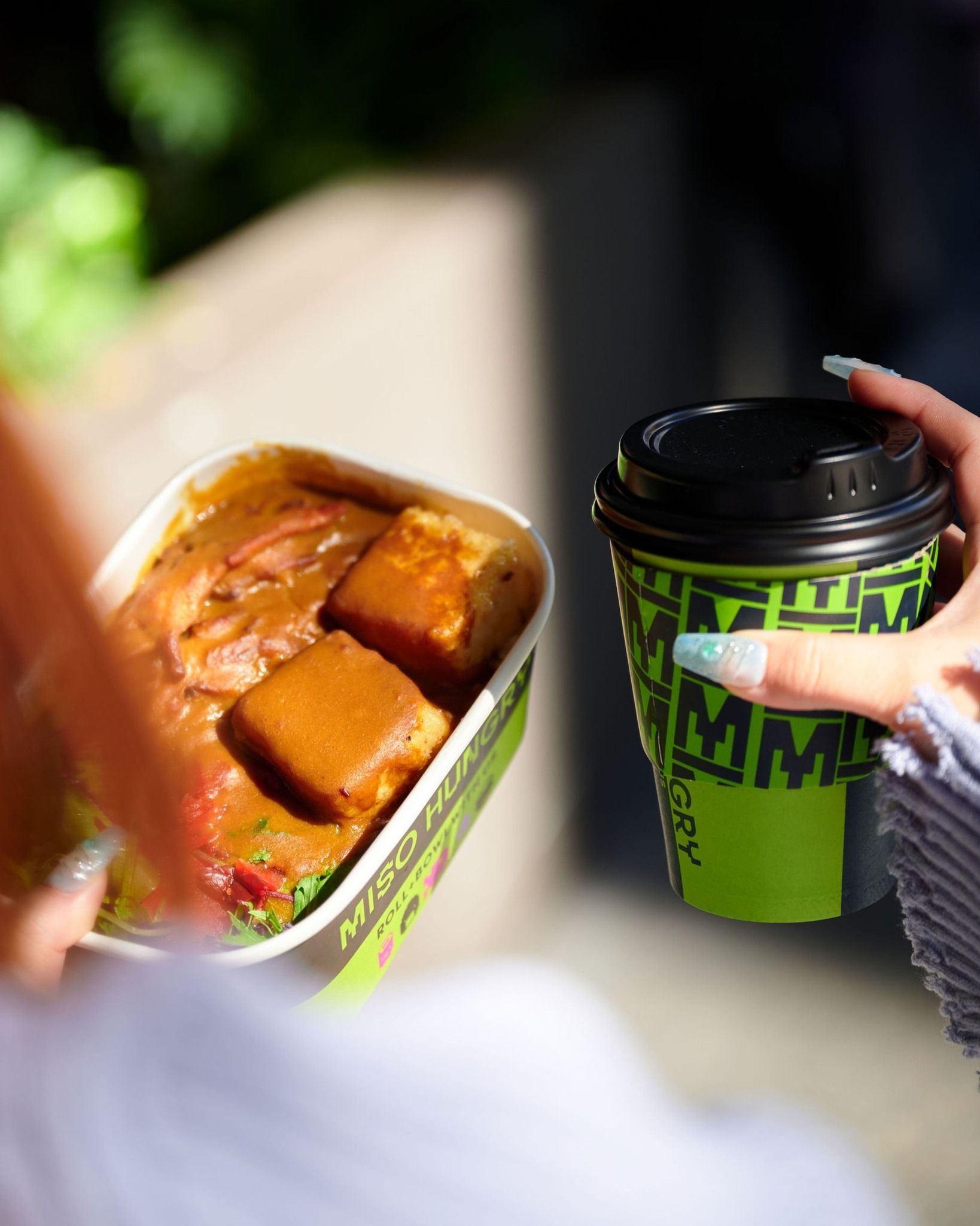 Miso Hungry - Hand holding a green cup beside an open takeout container with dumplings outdoors