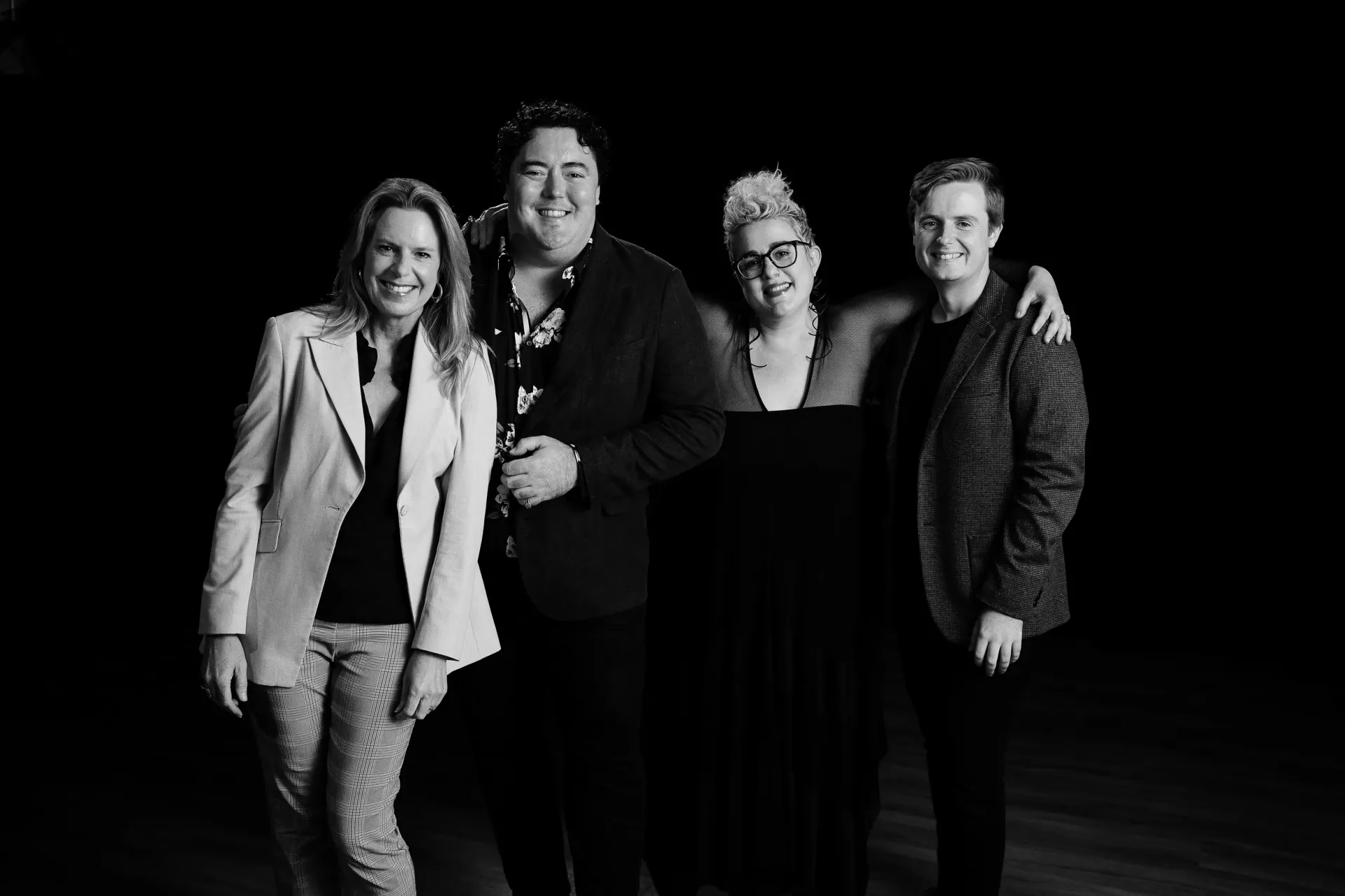 Australian Vocal Ensemble - Four people standing together against a black background, dressed in dark and light outfits, smiling at the camera.