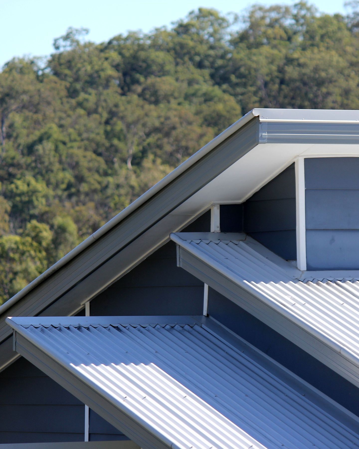 Elevation Metal Roofing - Blue corrugated metal roof with sloping gables against a wooded hillside background
