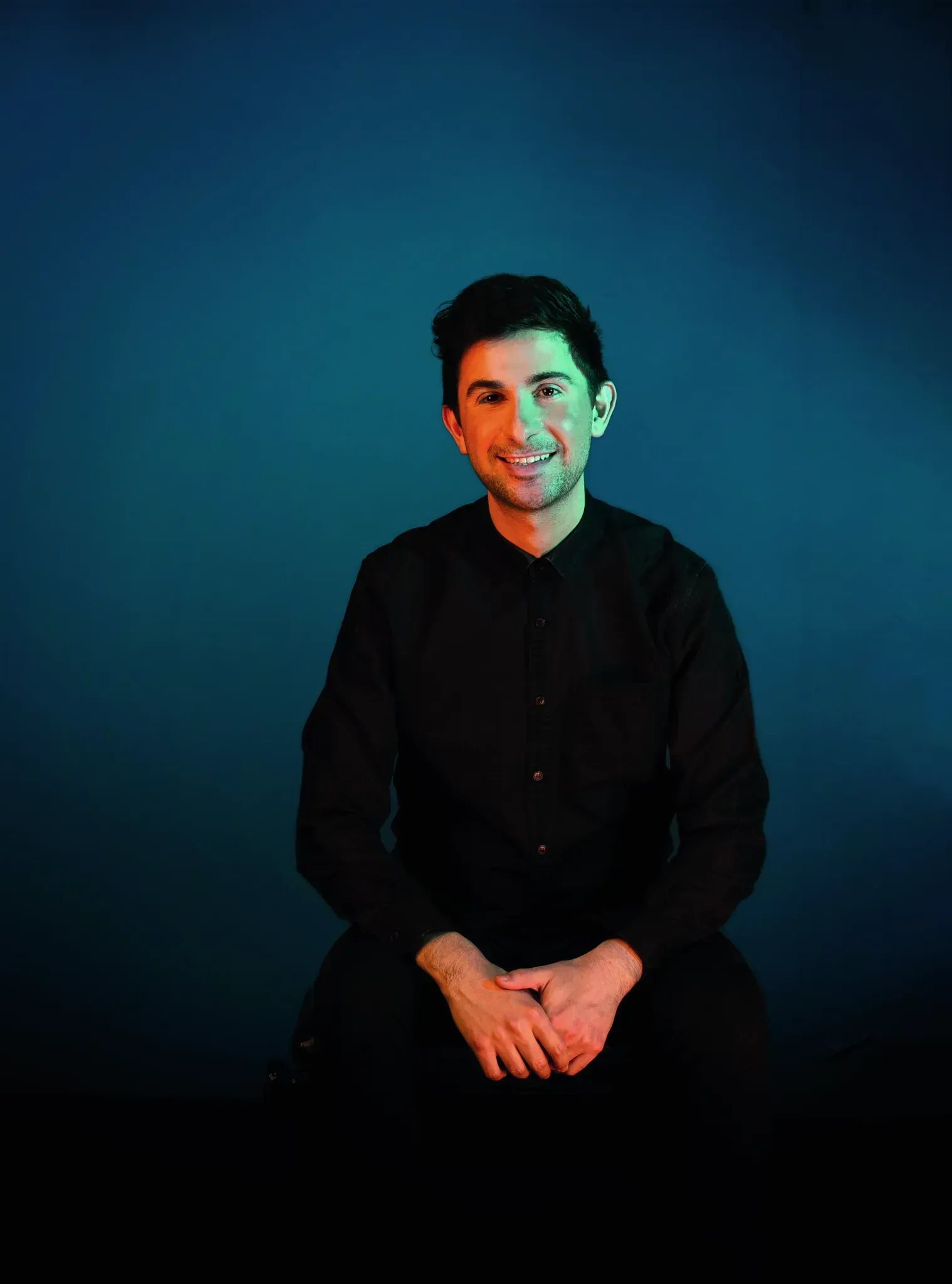 Alex Raineri - Person seated against a dark blue gradient backdrop, smiling, in a black shirt.
