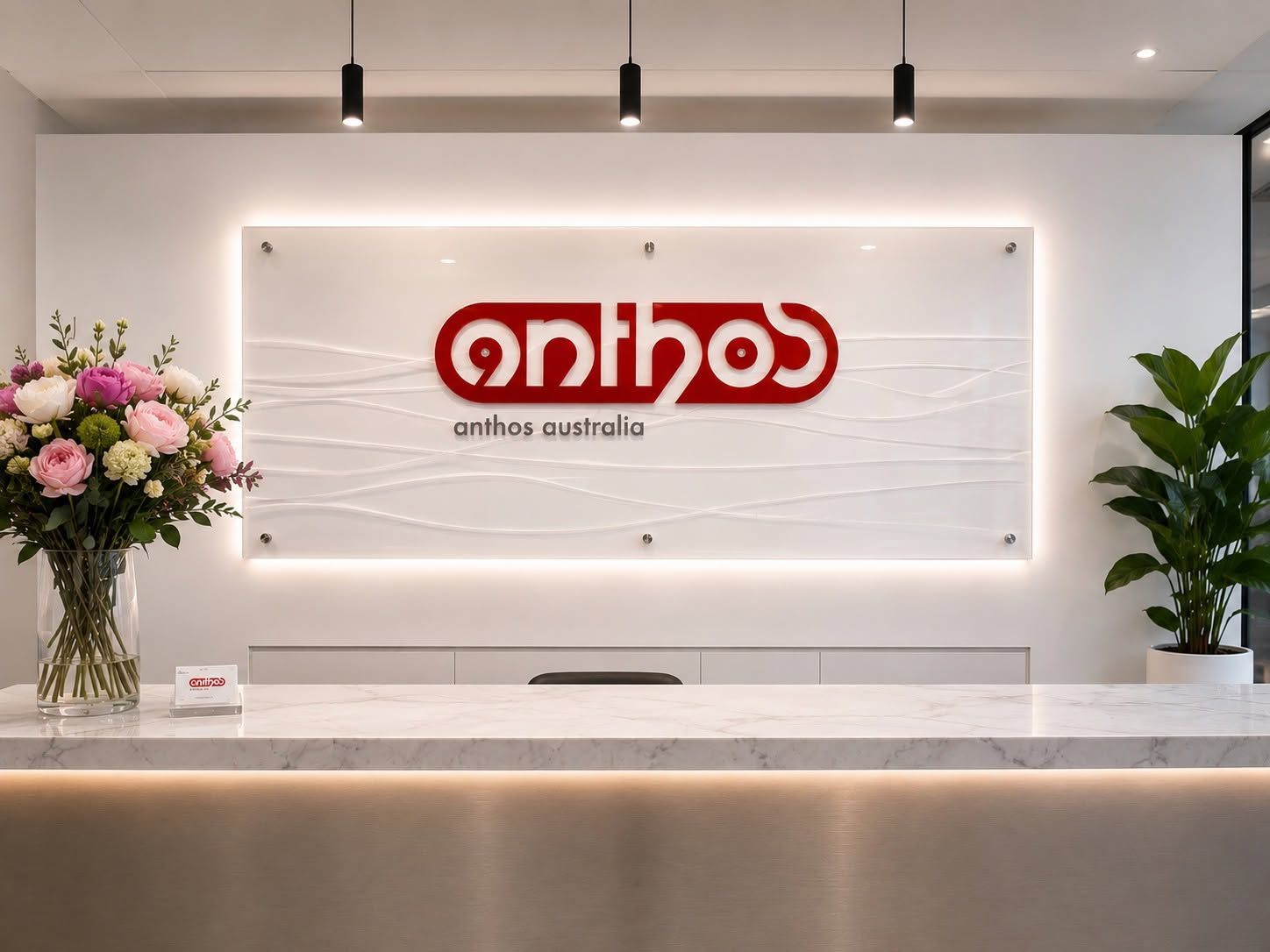 Anthos - Modern reception desk with backlit logo sign, white wall, flowers on the left, and tall plants on the right