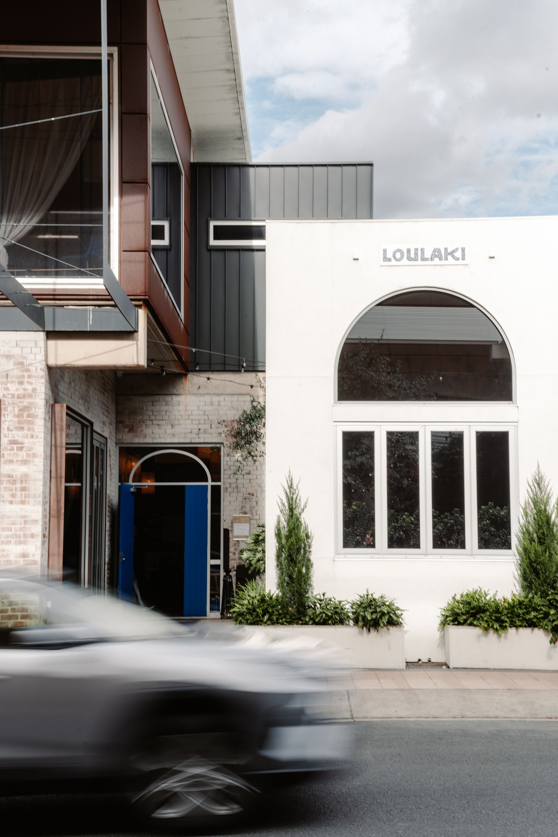 Loulaki white storefront with arched window and black trim on a city street, with a blurred car passing by