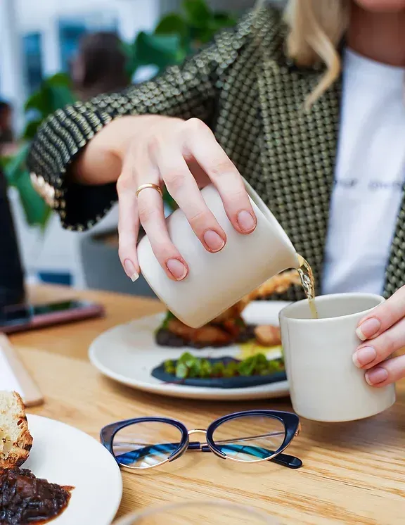 A woman is pouring coffee into a cup while sitting at a table.