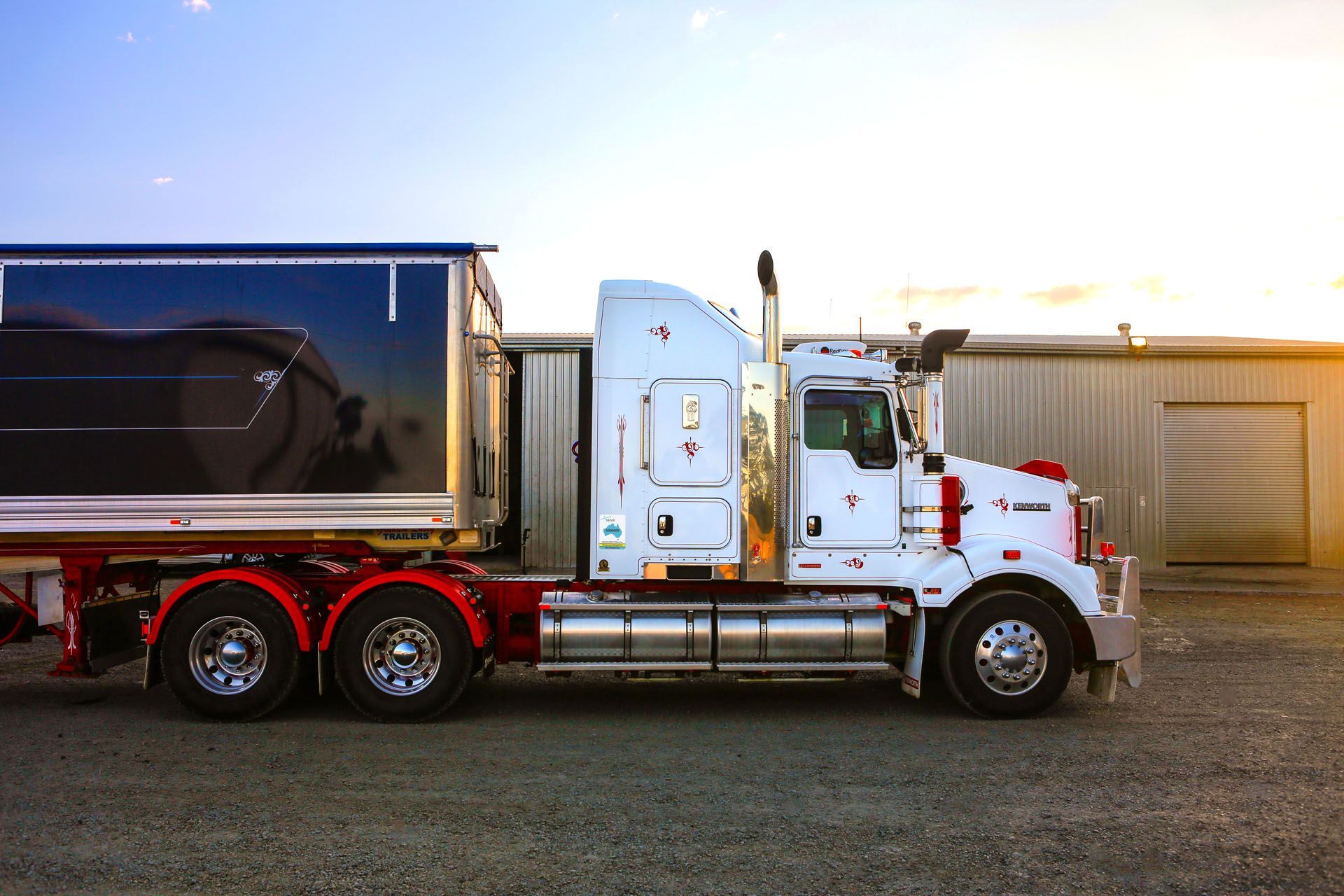 NHVR - White semi truck with black trailer parked on gravel in front of a warehouse at sunset