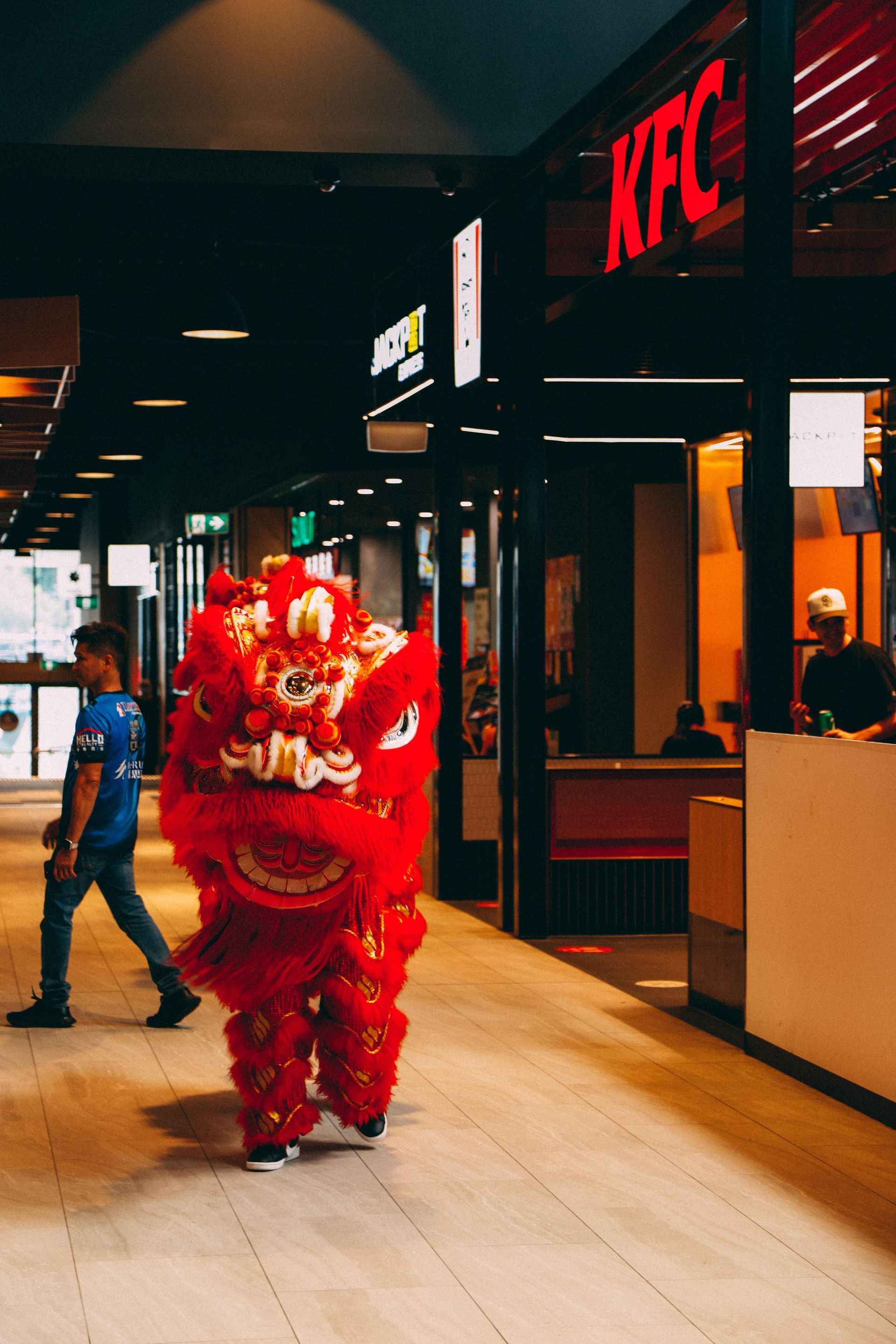 A person in a red lion costume is walking in front of a kfc restaurant.