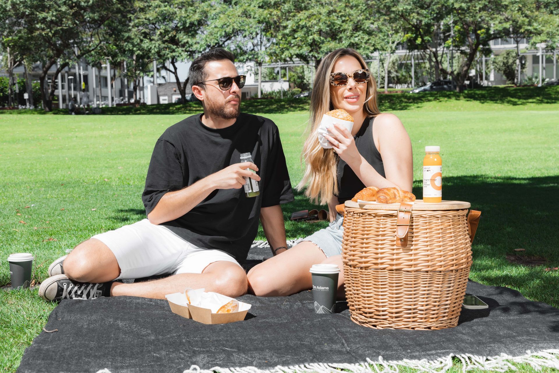 A man and a woman are sitting on a blanket in a park with a picnic basket.