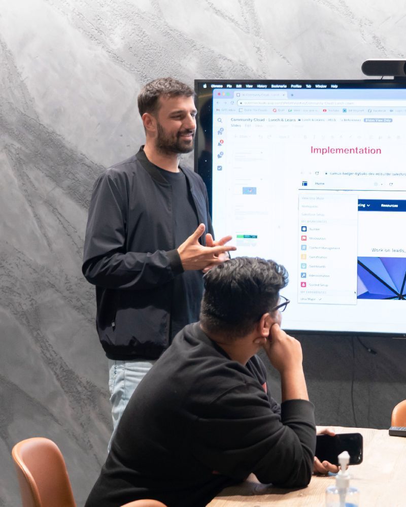 Out in the Clouds - Two people in a meeting room, one presenting beside a large screen with a website displayed.