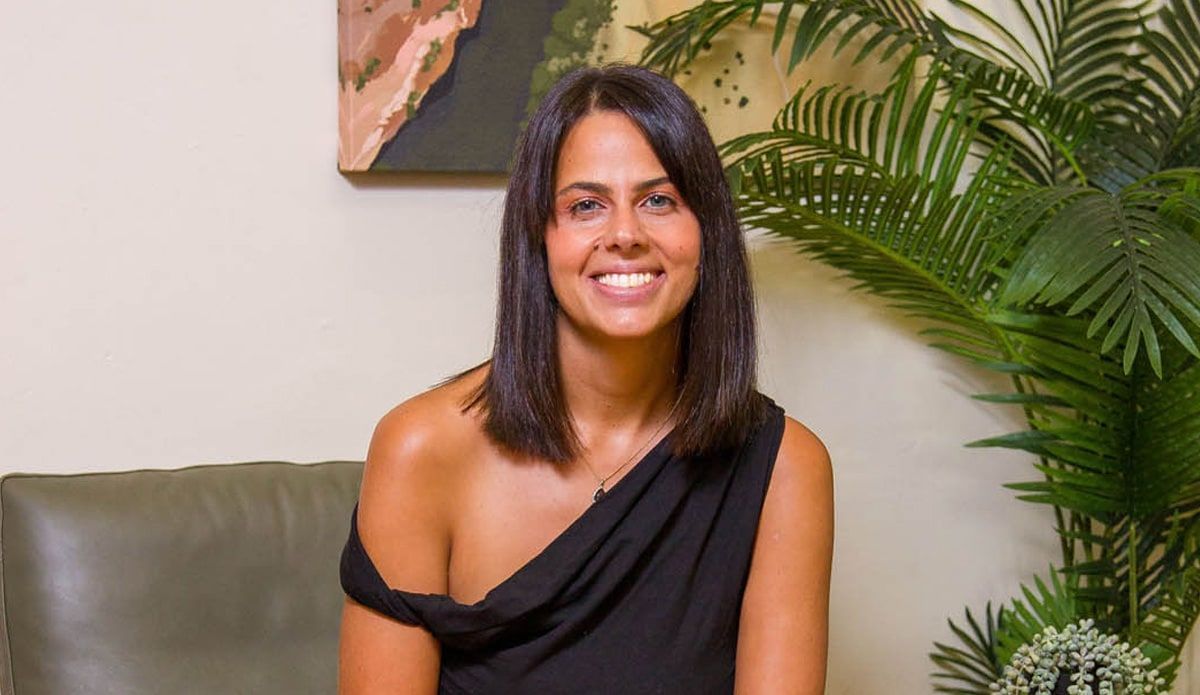 Sharne Consulting - Woman smiling on a sofa indoors beside a large green plant.