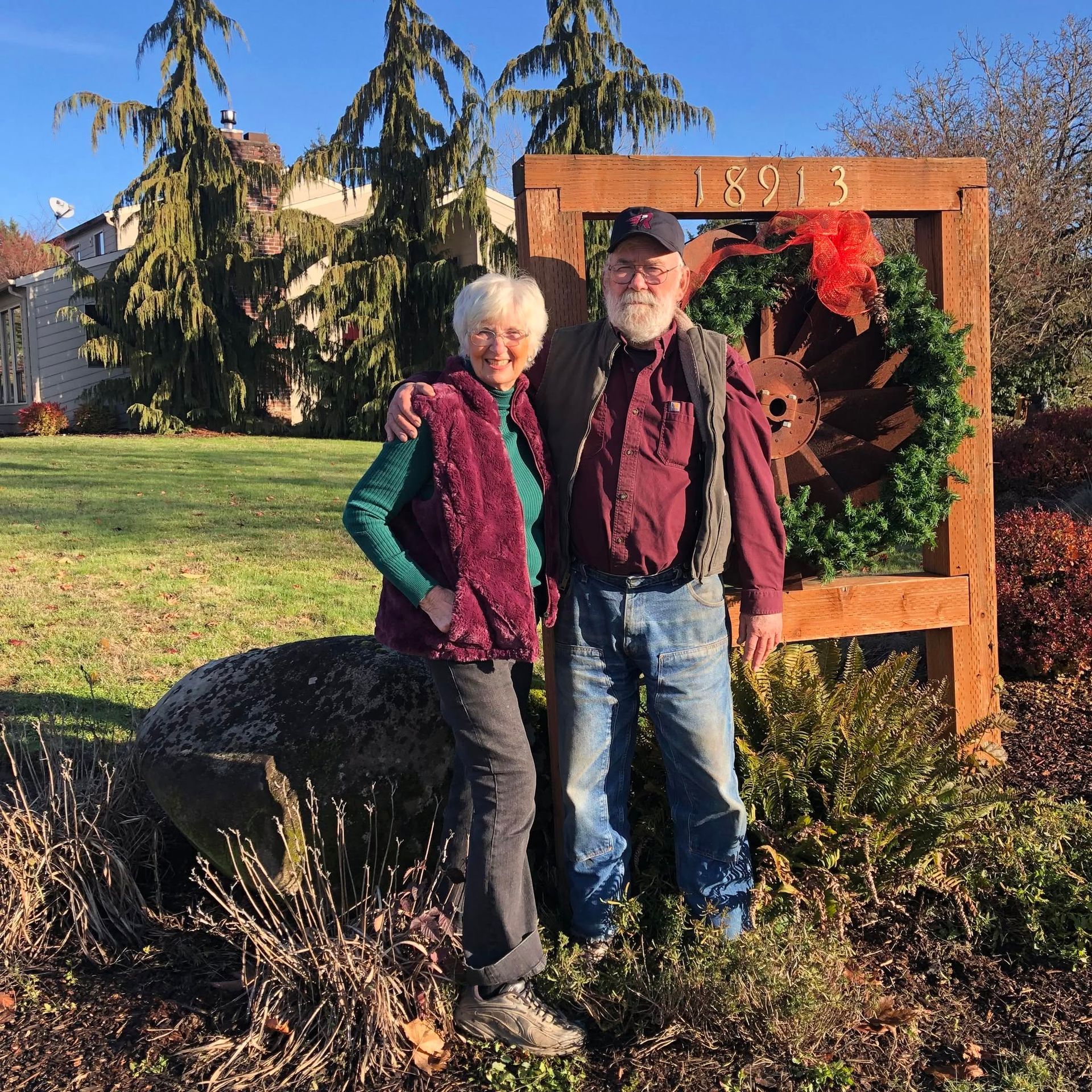 Two people standing outdoors beside a large wooden gear sculpture in a sunny garden