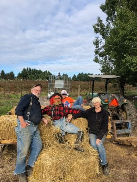 Four people posing on hay bales in a pumpkin patch beside a tractor under a cloudy sky