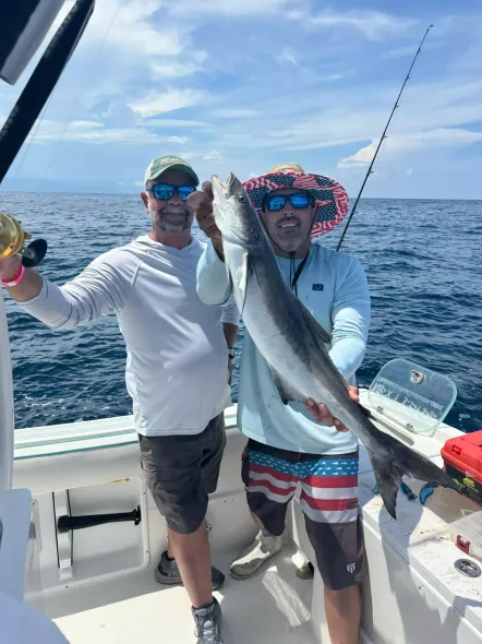 Two men on a boat hold up a large fish, with fishing rods and open ocean behind them.