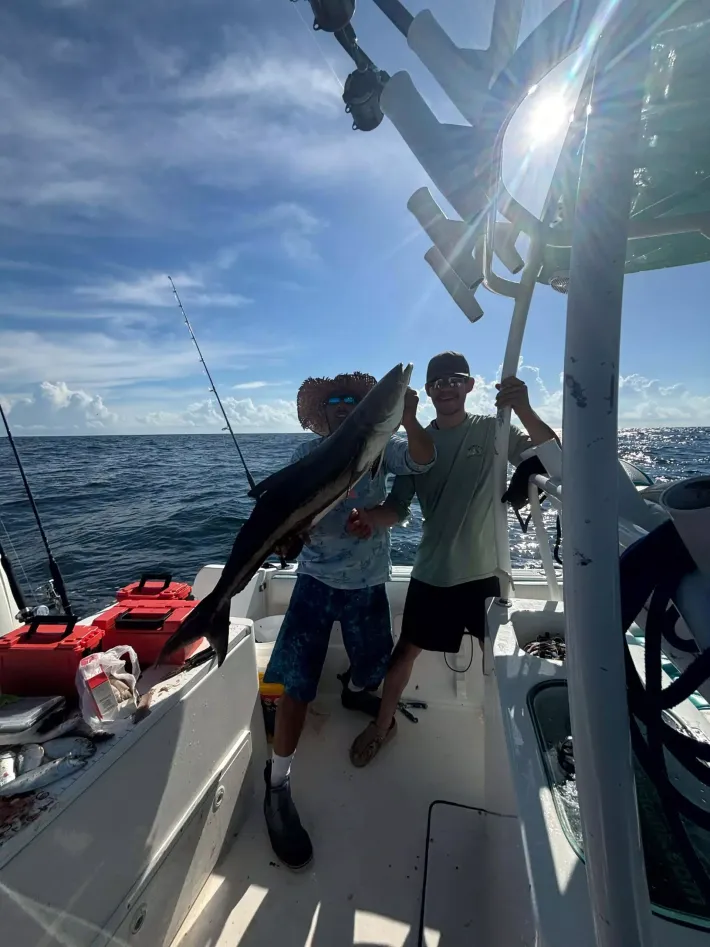 Two people on a fishing boat holding up a large fish on a sunny open sea.