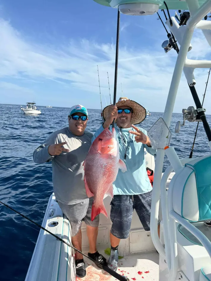 Two anglers on a boat hold a large red fish over the ocean, with another boat in the distance.
