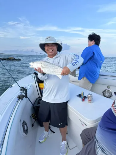 Two people fishing on a boat; one holds up a caught fish under a bright blue sky.