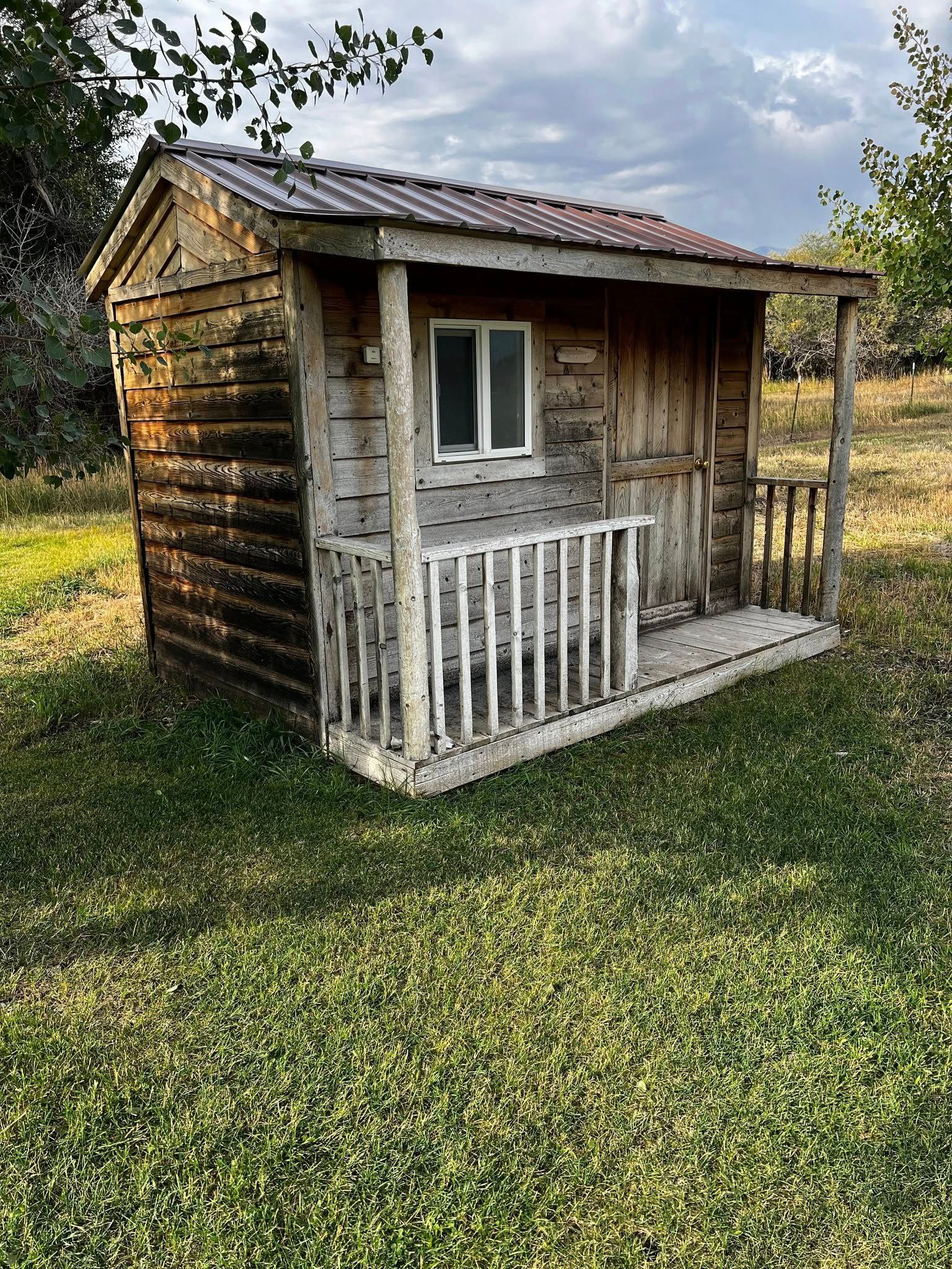 Weathered wooden shed with a small porch, set in grassy field under a cloudy sky.