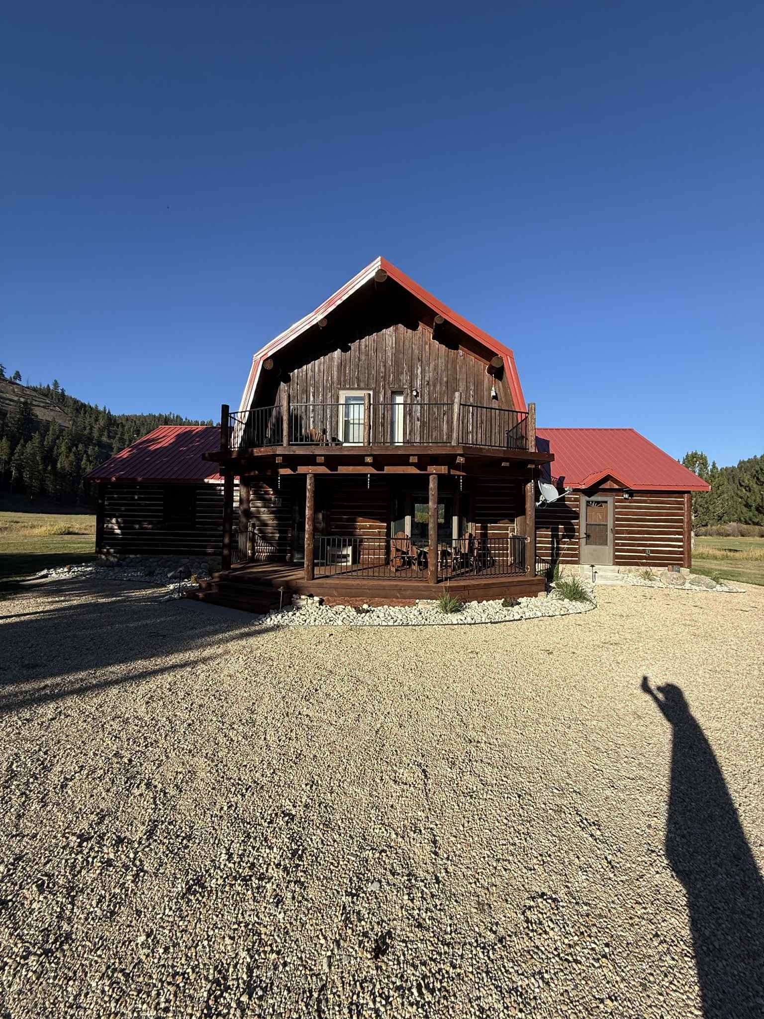 Rustic log cabin with red roof and wraparound porch on a gravel lot under a blue sky.
