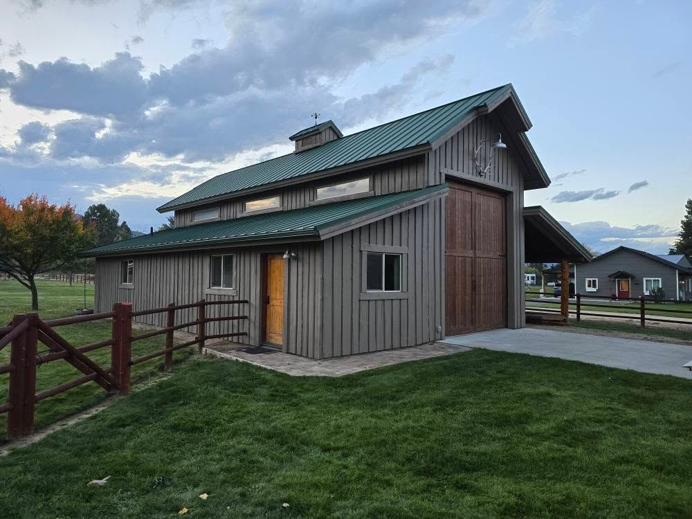 Wooden barn with green roof, open door, and adjacent carport in a grassy yard under a cloudy sky.