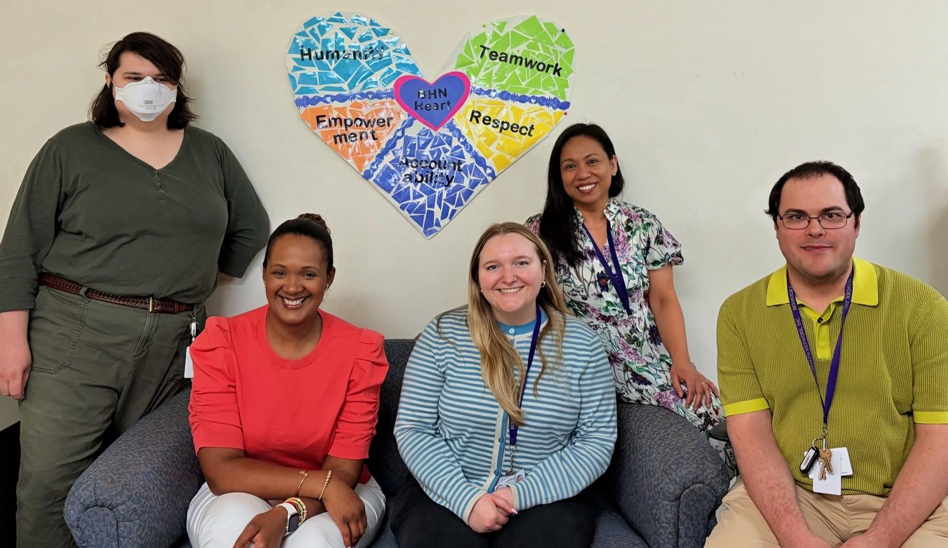 Five BHn Employees sitting together next a heart shaped art peice they created displaying BHN Heart Values.