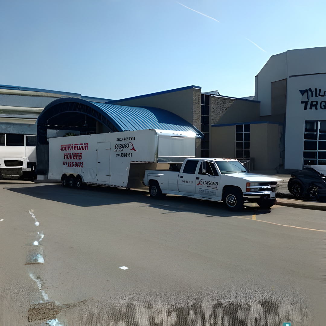 A white pickup truck with a large, white gooseneck trailer parked in front of a building with a blue curved roof.
