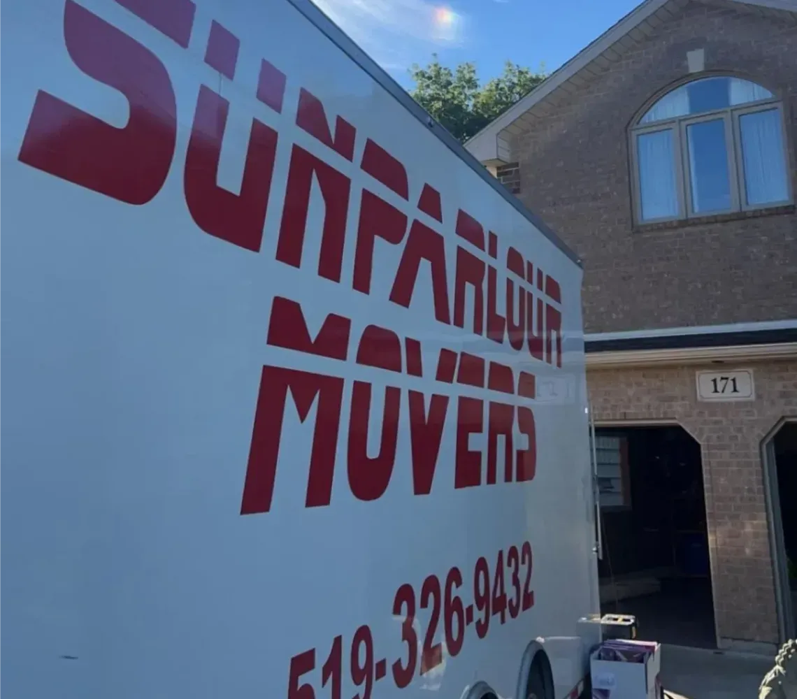 A Sunparlour Movers trailer parked in a residential driveway in front of a brick house with a garage.