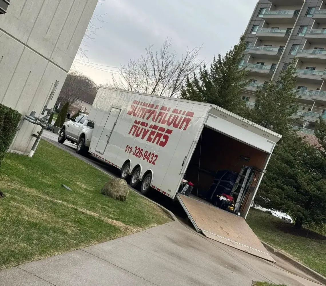 A white cargo trailer with a lowered rear ramp sits on a grassy area next to an apartment building.