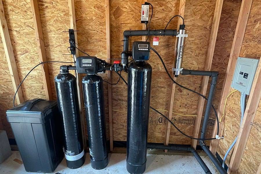 Water softener and filtration system with black tanks and pipes installed against an unfinished wall.