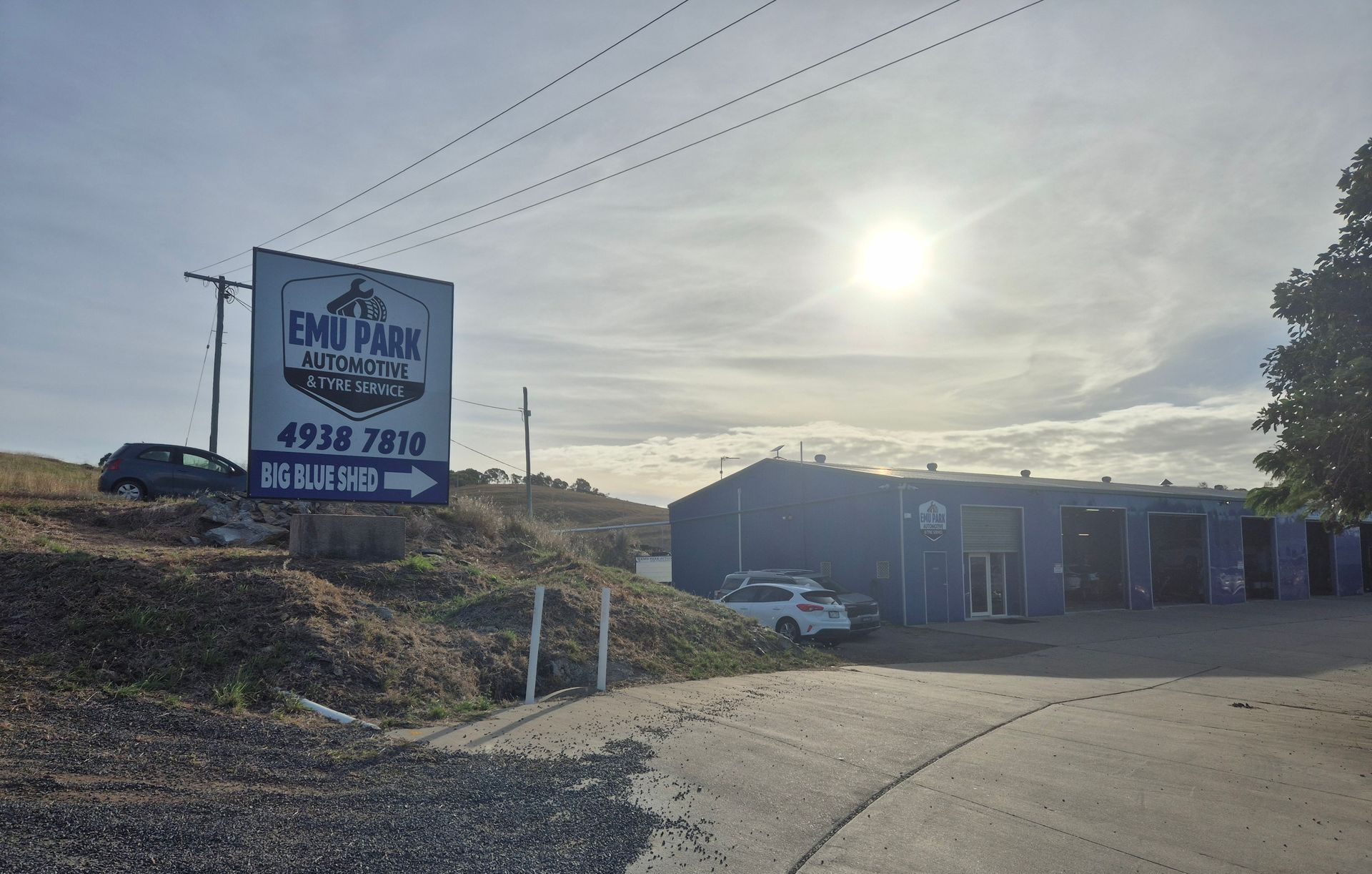 Roadside building with an ENMU sign, utility lines, and bright sun over a paved lot.  — Emu Park Automotive and Tyre Service in Emu Park, QLD