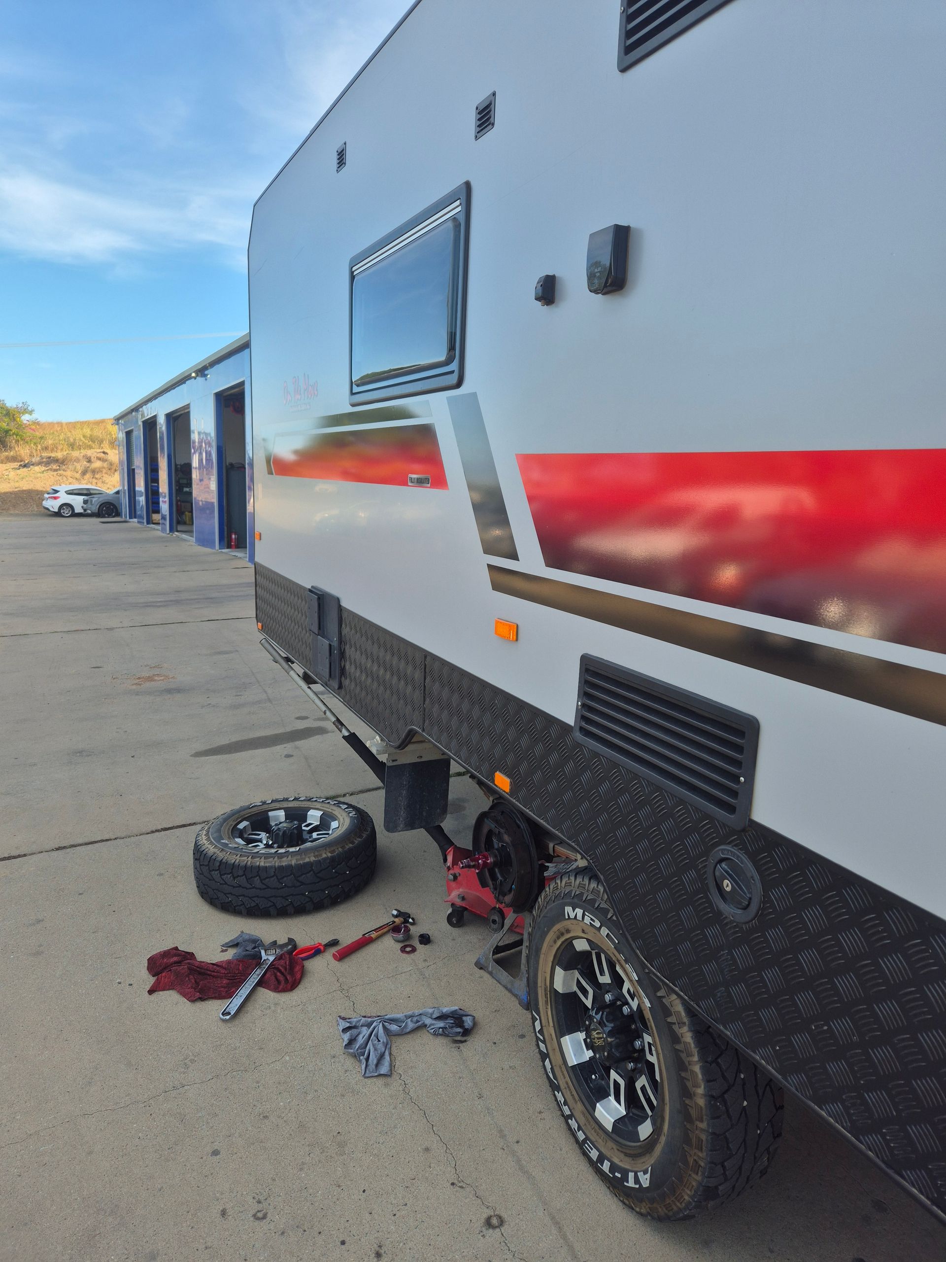 White RV trailer with red stripe parked on pavement; detached tire and tools lie beside the wheel. — Emu Park Automotive and Tyre Service in Emu Park, QLD
