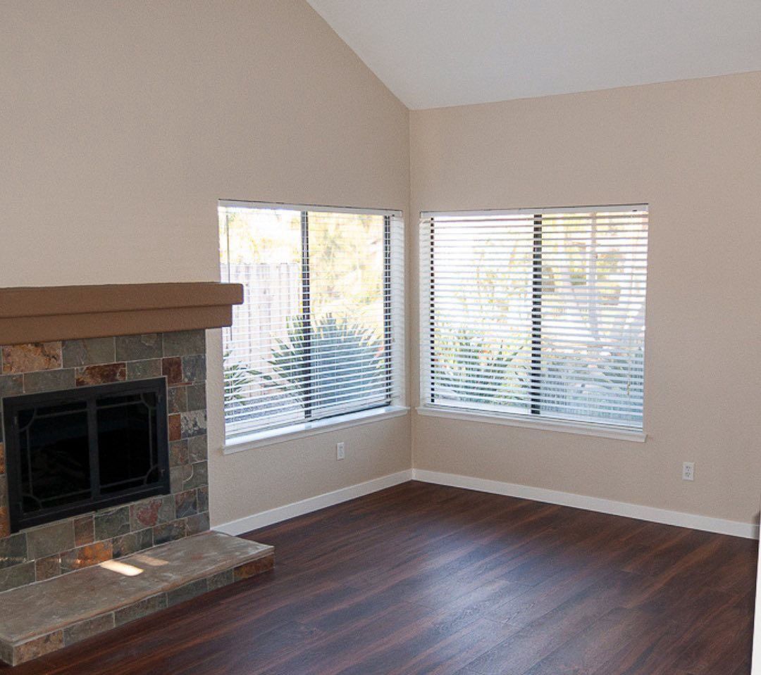 Empty living room with a stone fireplace, dark wood floor, and two bright corner windows