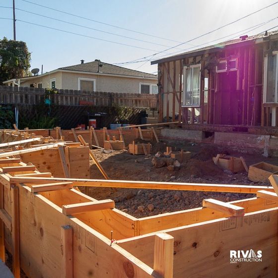 Wooden house foundation framing at a construction site beside a partially built home in bright sunlight