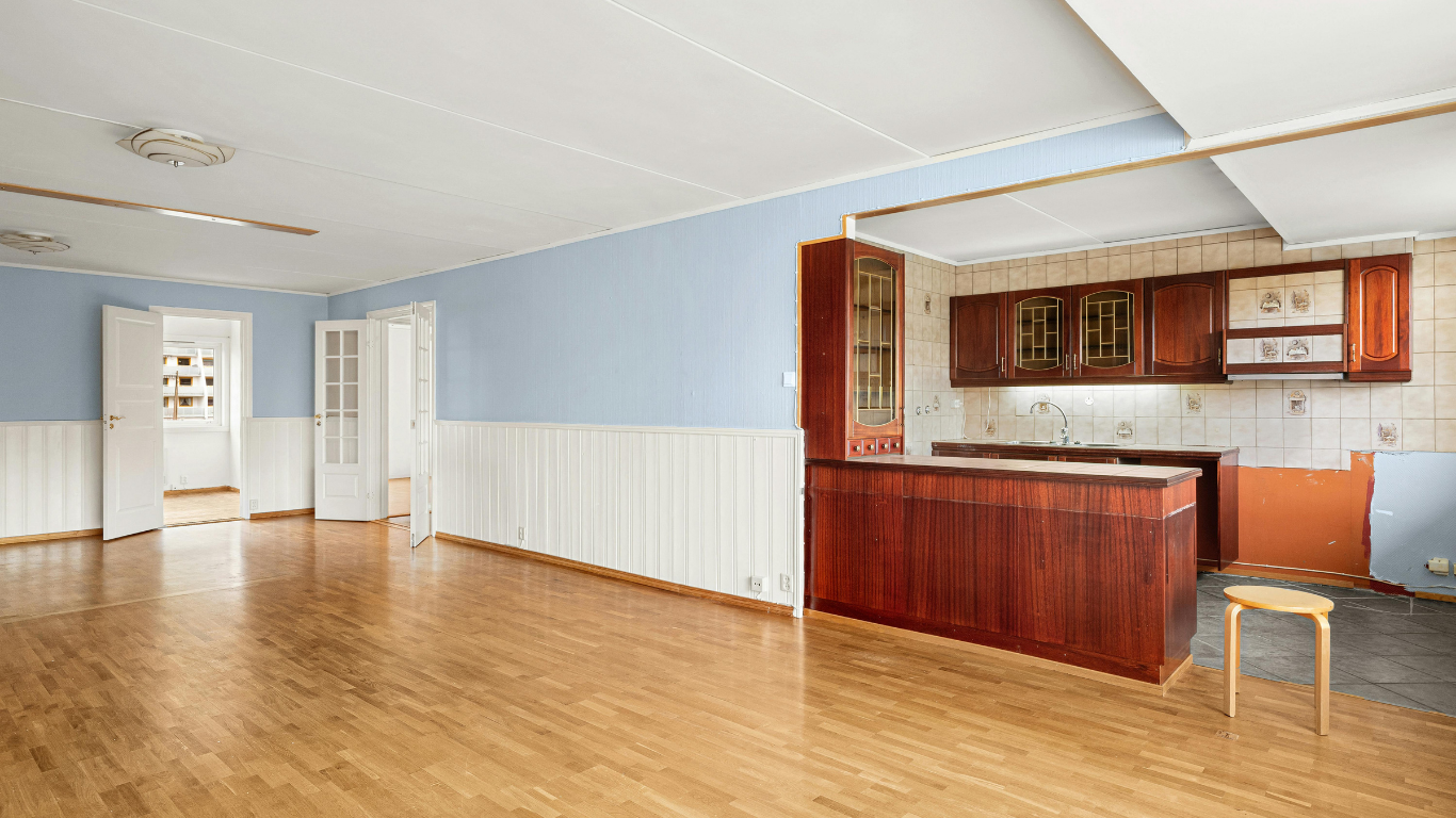 Empty living room with light blue walls, hardwood floor, and a small kitchen with wooden cabinets and bar counter.