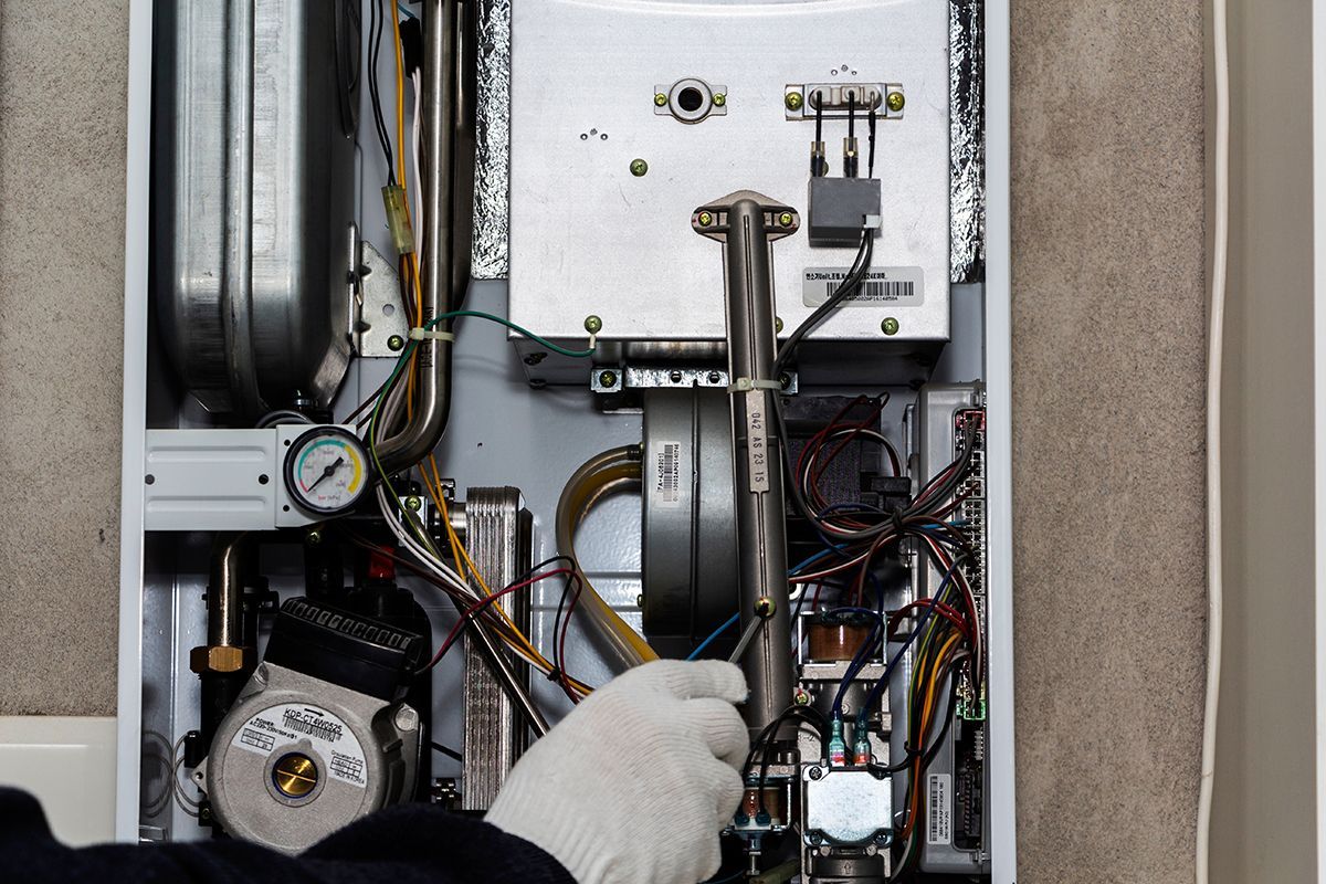 A person wearing a white glove works on the internal components of an open wall-mounted boiler.