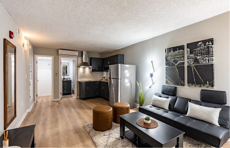 Modern Studio Apartments In Nashville, TN - Riverwalk 803 - Living Room With Vinyl Plank Flooring, Area Rug, Leather Couch, Coffee Table, Leather Ottoman Stools, And View Of Kitchen Area.
