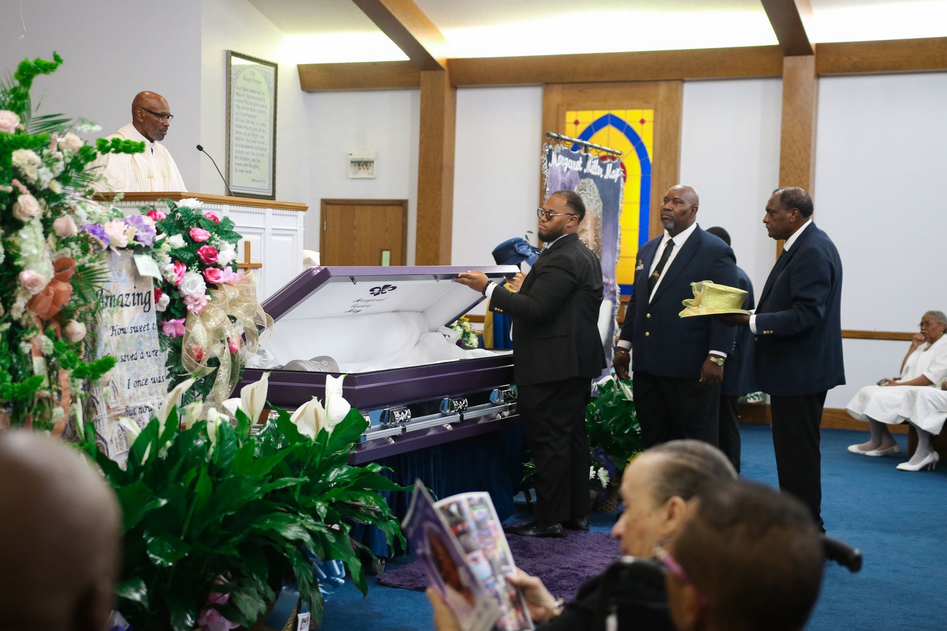 Funeral scene with floral casket and mourners standing beside it in a church-like setting