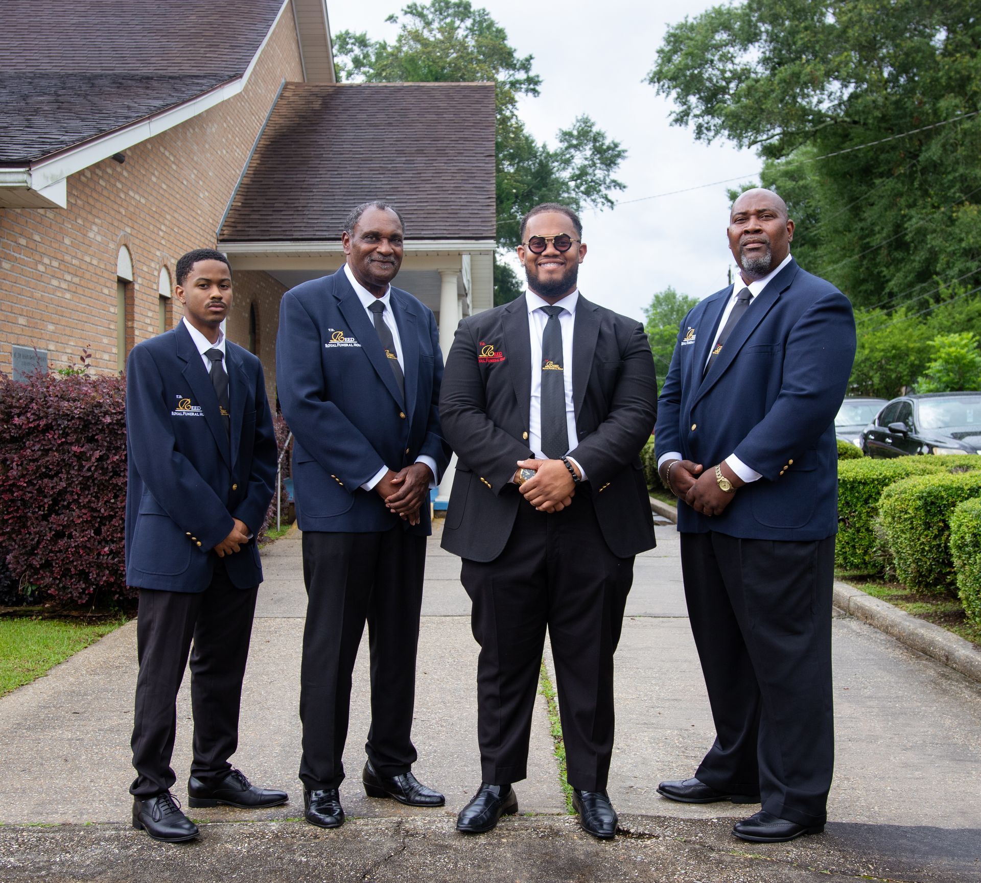 Four men in dark suits standing together outside a building on a driveway.