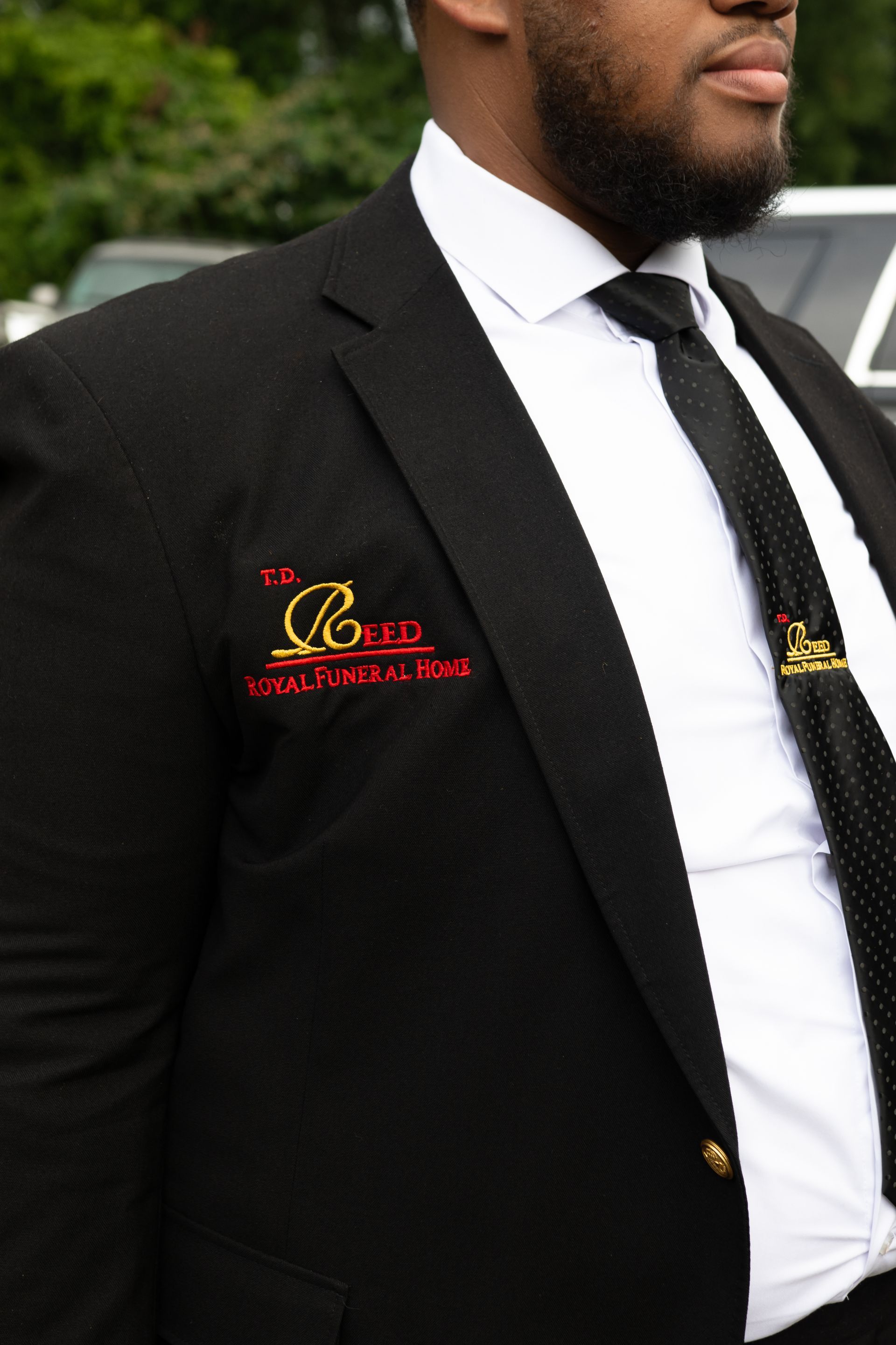 Man in a black suit and tie with a red logo on the jacket, standing outdoors.