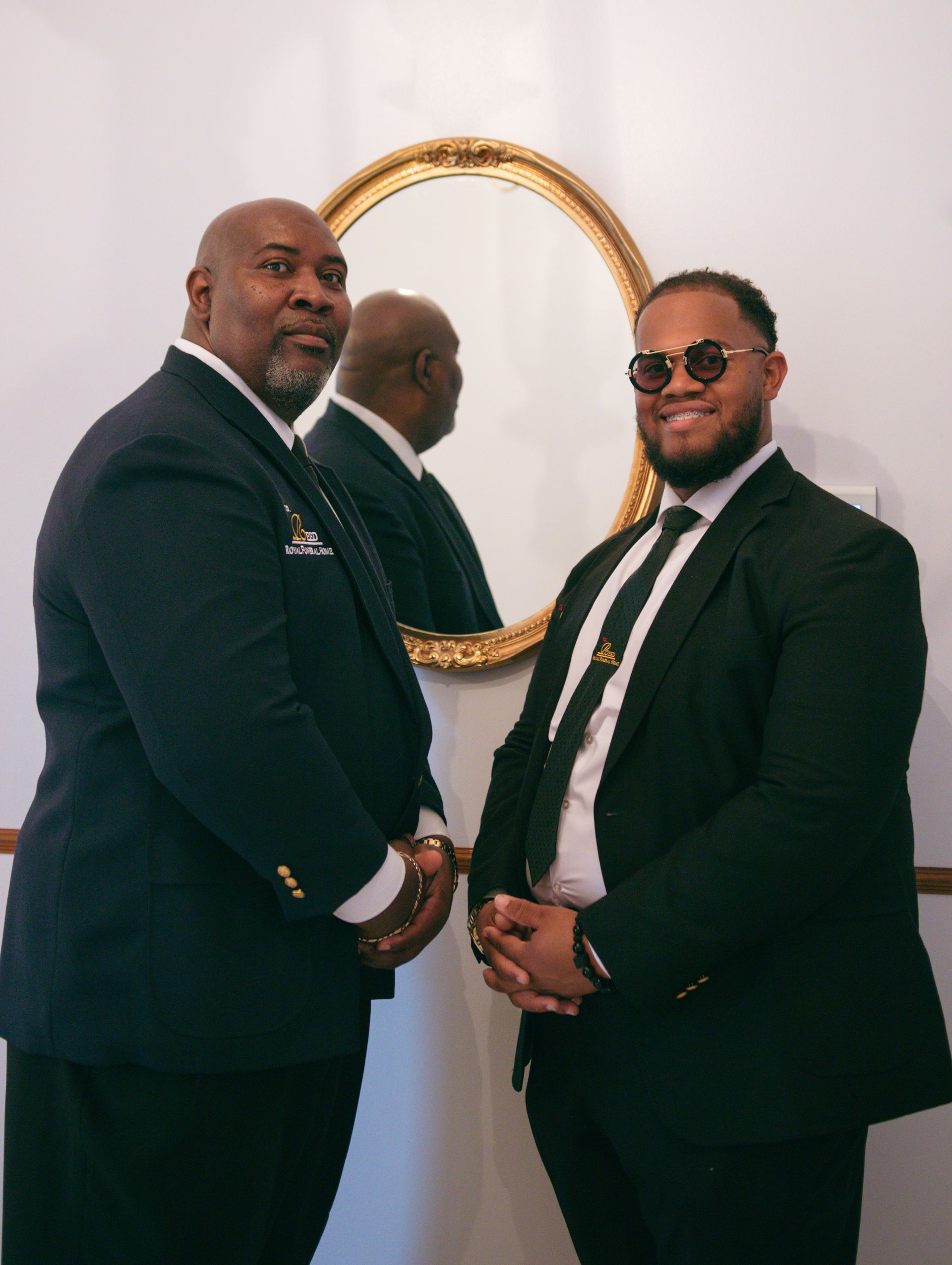 Two men in dark suits stand beside a gold-framed mirror in a white room.