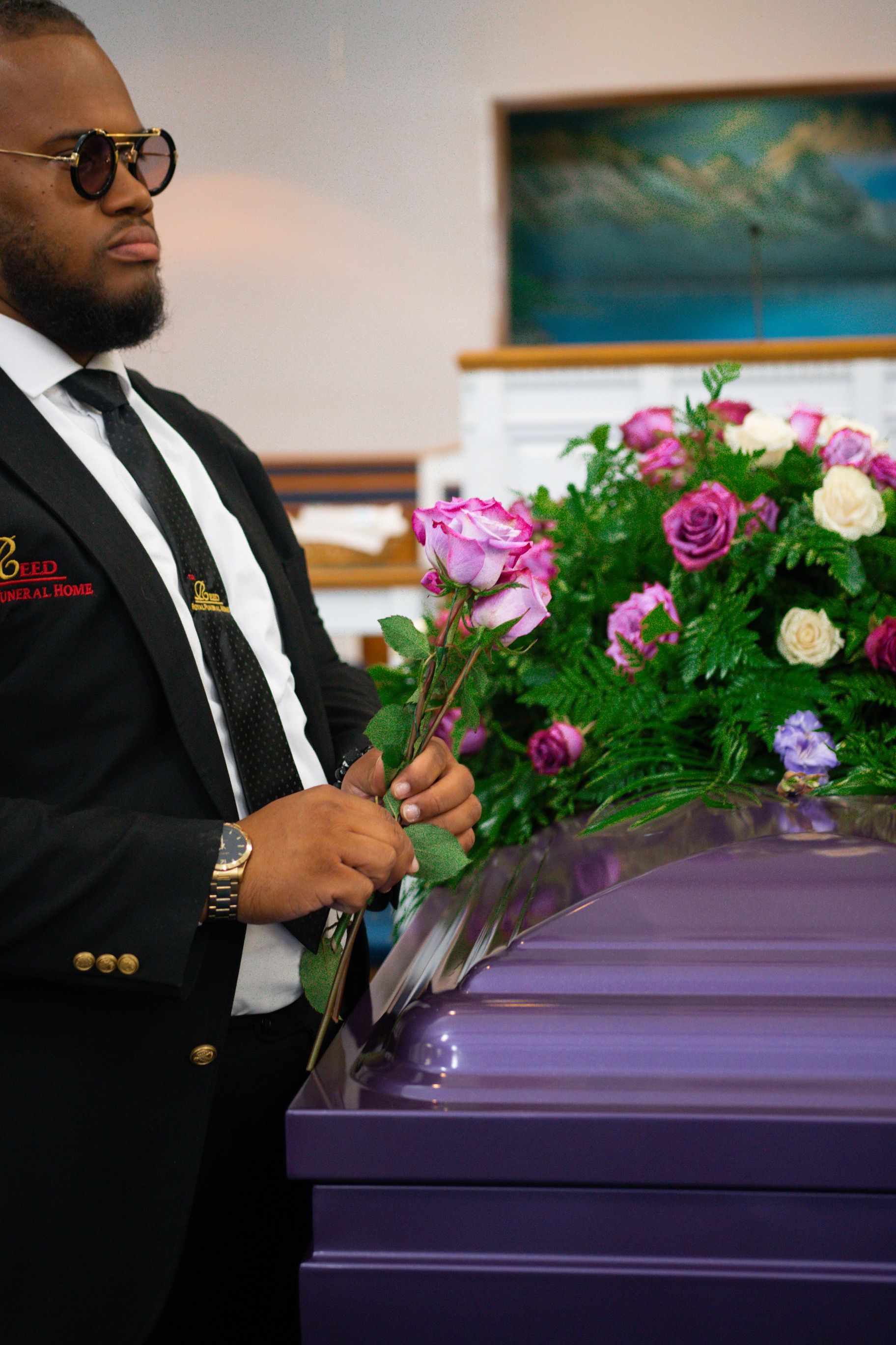 Man in black suit holding a flower at a purple casket with pink and purple flowers