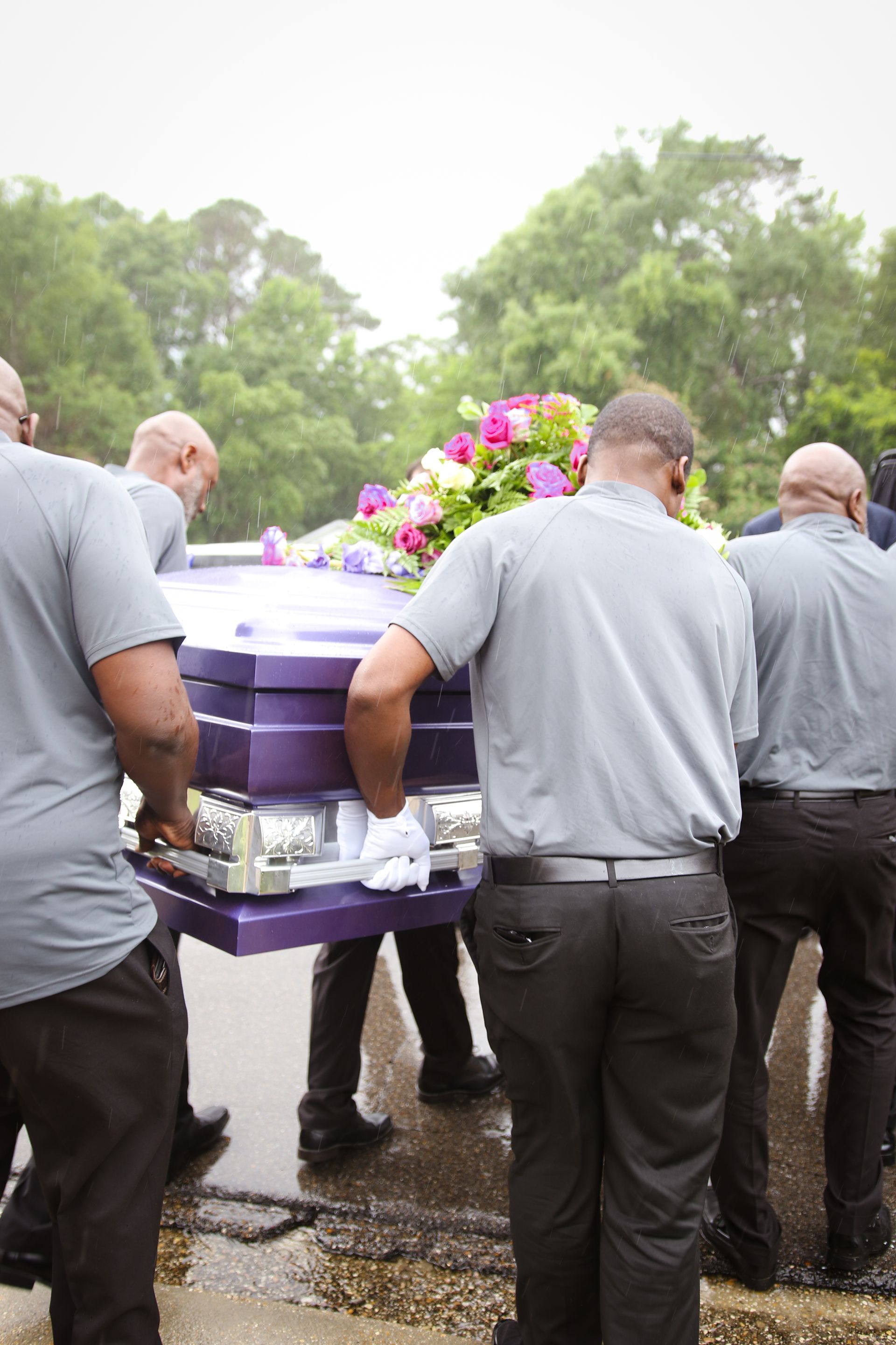 Mourners carrying a purple casket with flowers at a rainy outdoor funeral procession