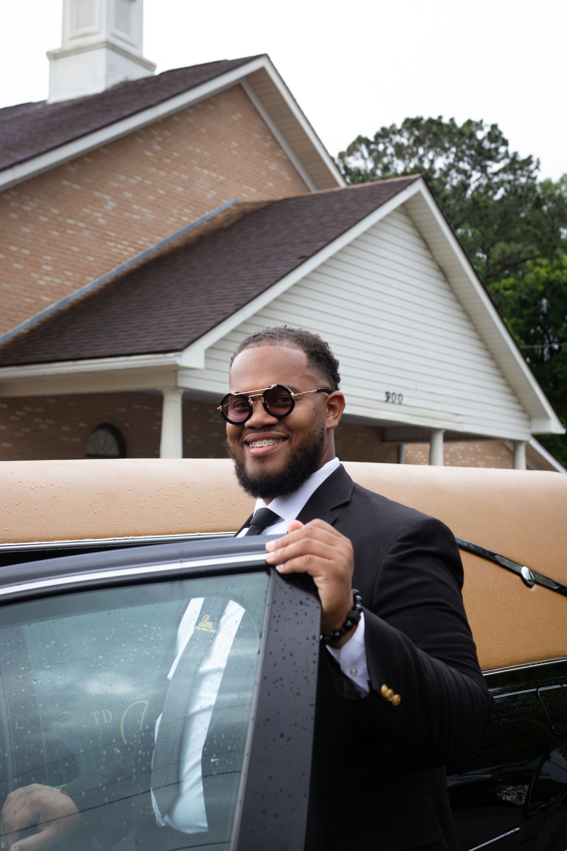 Smiling man in a black suit standing by a car in front of a house