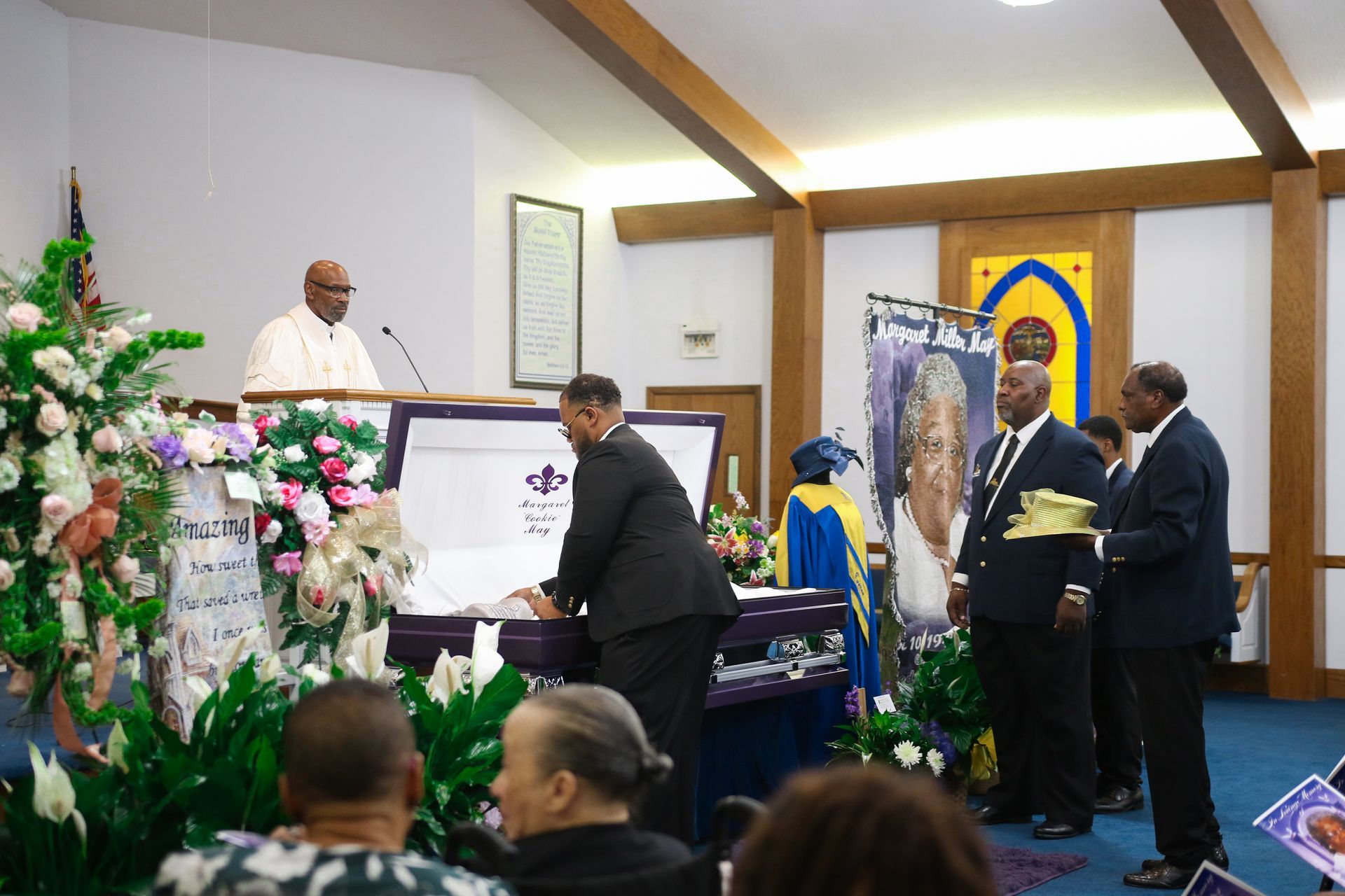 Mourners at a church service beside a flower-covered casket and speaker at the pulpit