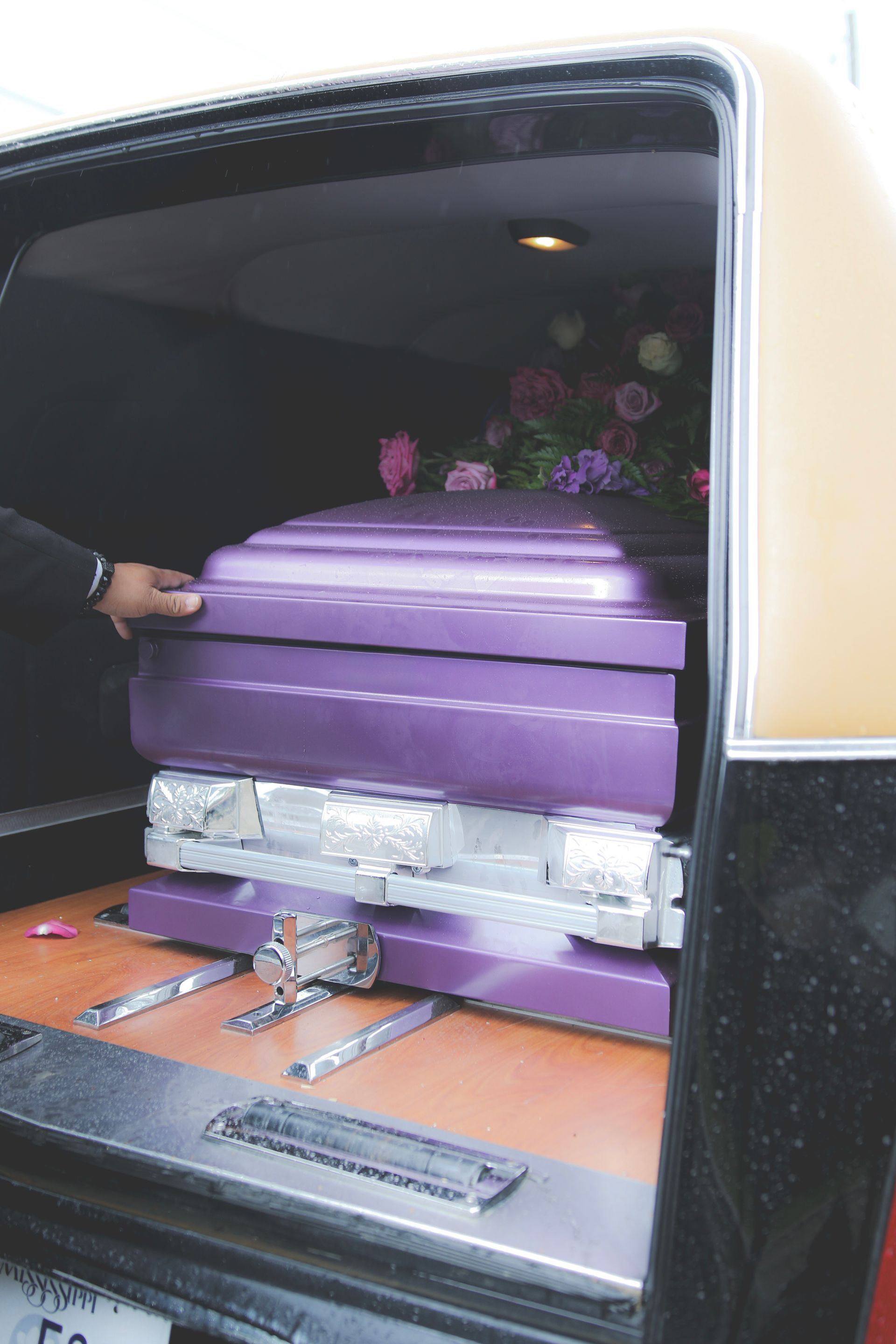 Purple coffin being slid into a hearse, with flowers visible inside.