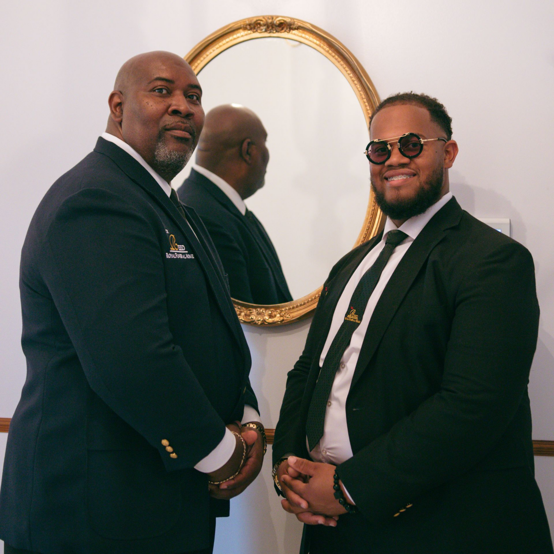 Two men in dark suits posing indoors beside a gold-framed mirror.