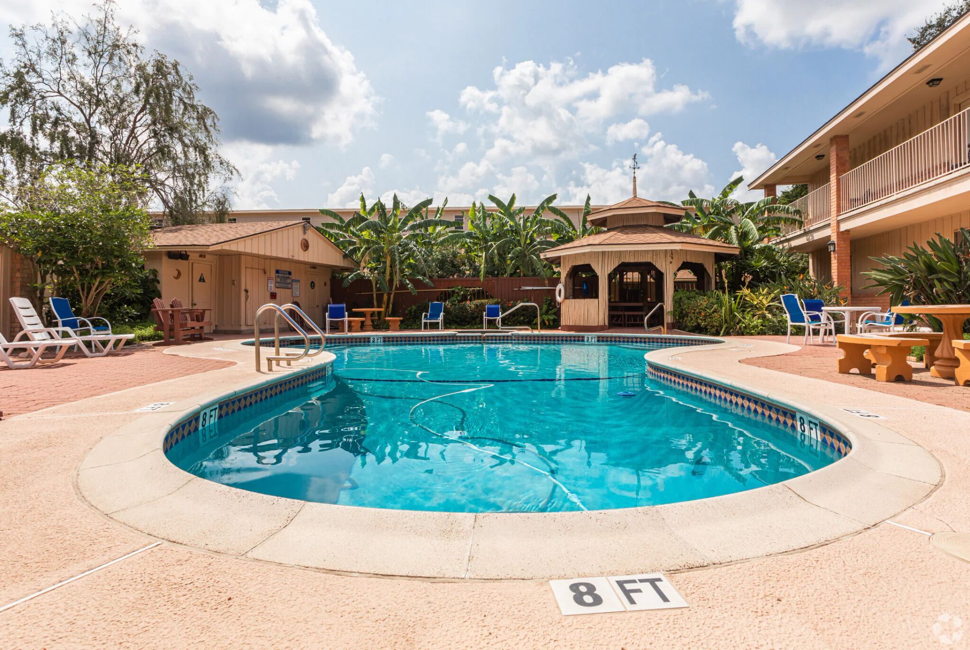 Swimming pool surrounded by a beige patio, a gazebo, and buildings. Sunny day with clouds.
