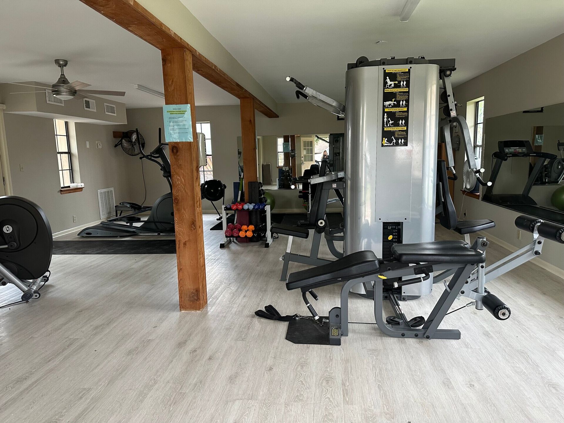 Gym interior with exercise equipment: weights, treadmill, and multi-gym machine. Wooden support beams.