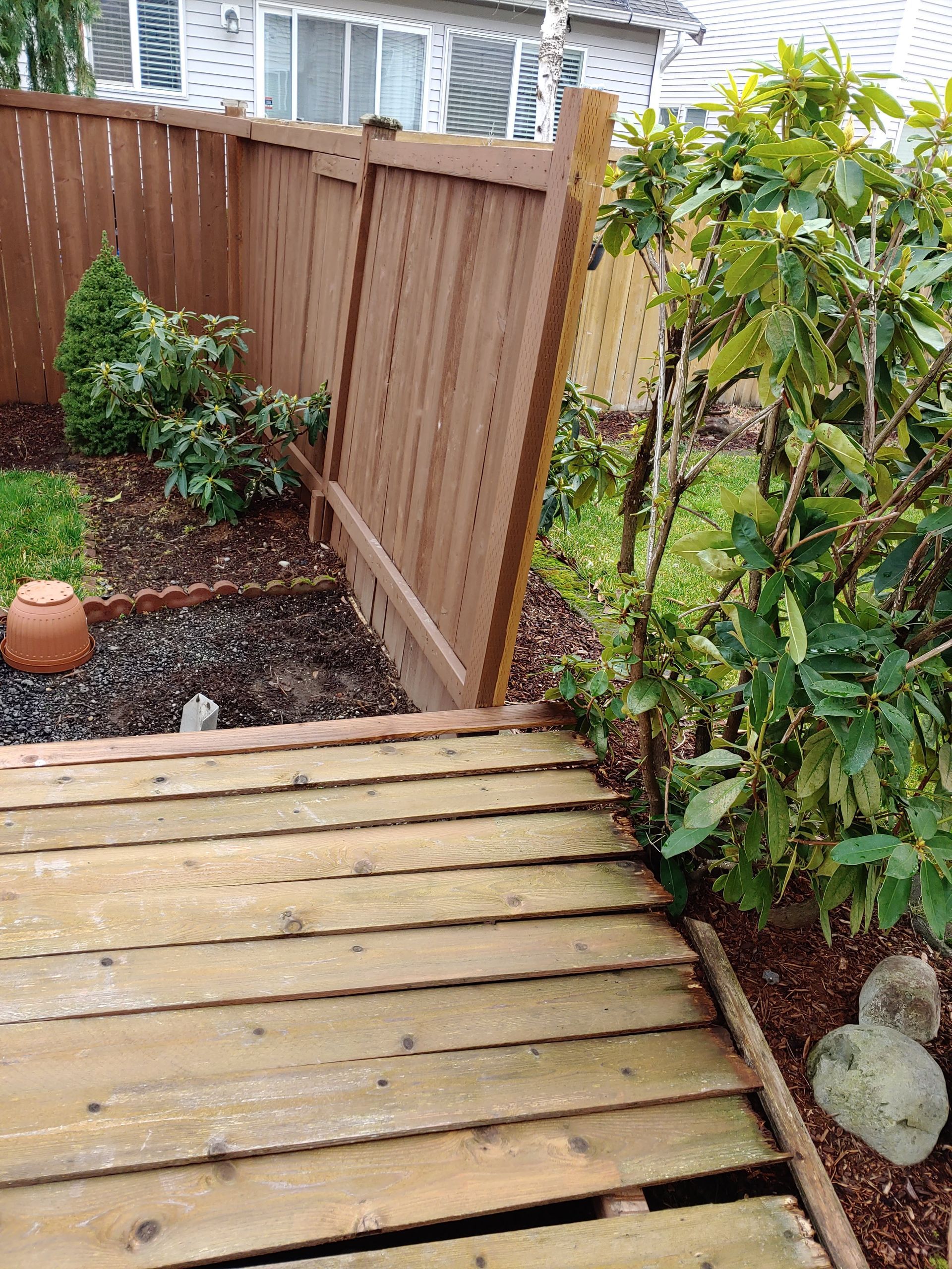 A wooden deck platform sits in a yard next to a brown wooden fence, garden beds, and a leafy green shrub.