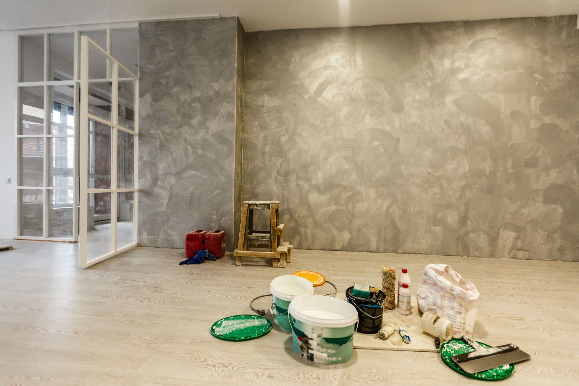 Paint supplies and tools on the floor of a room with a textured grey concrete wall and a white grid-pattern door.